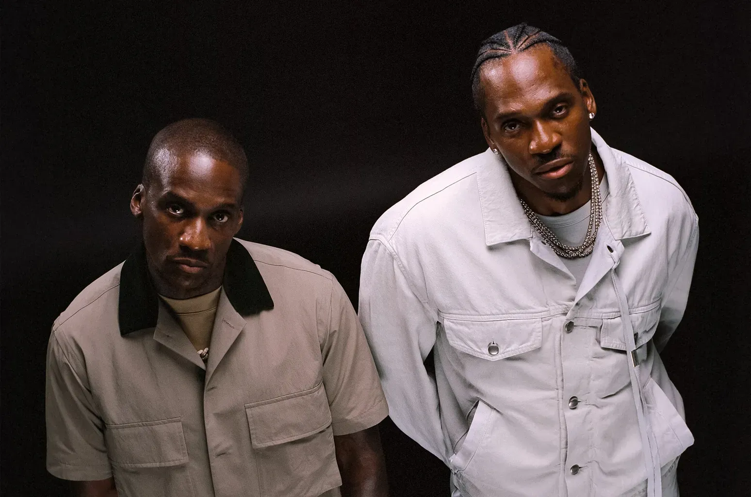 Two men pose against a black background, one in beige and one in white, both looking at the camera.