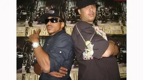 Two men in dark shirts and hats stand back-to-back in front of a wall of cash.
