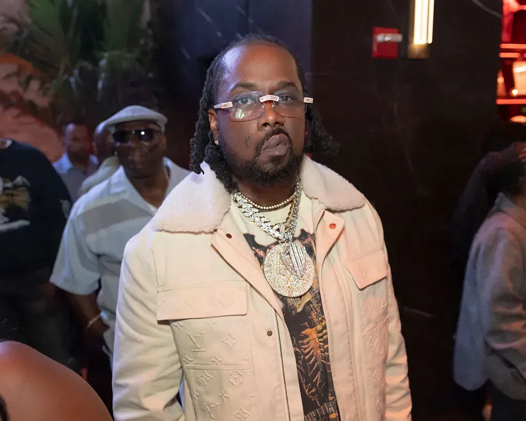 Man in beige jacket with layered necklaces stands in a dimly lit nightclub or event space.