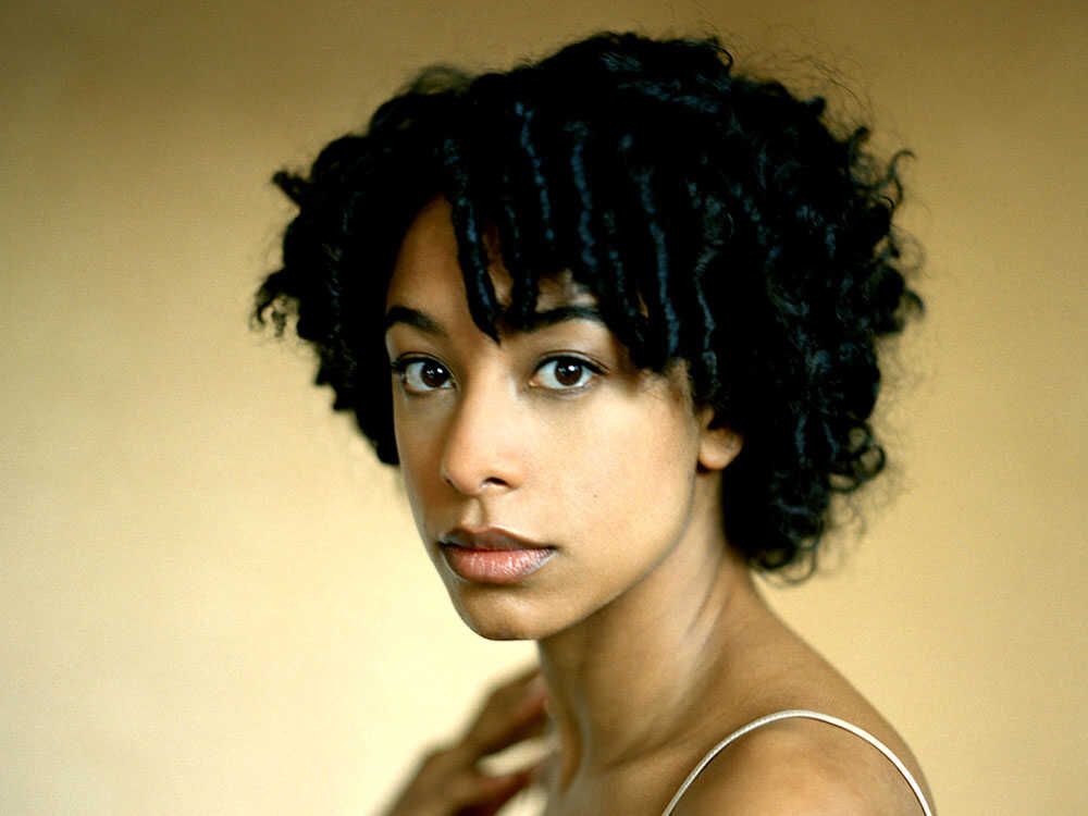 Portrait of a person with curly dark hair against a warm beige background, looking at the camera.