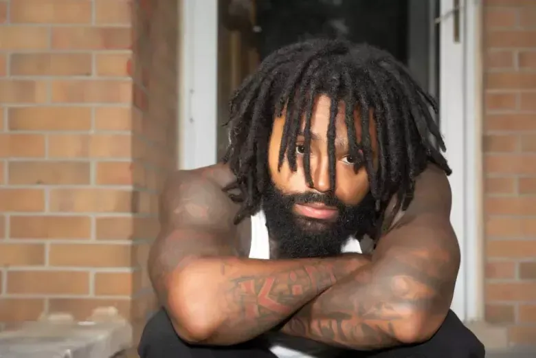 Man with dreadlocks and beard resting his arms on a table against a brick wall, looking at the camera.