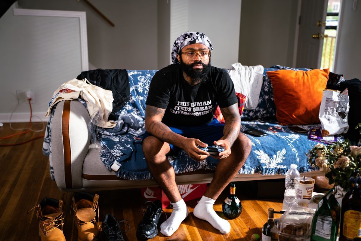 Person sitting on a couch playing a video game, with a blue blanket and orange pillow in a living room.