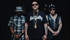 Three people posing against a dark background, wearing sunglasses, hats, and dark streetwear.