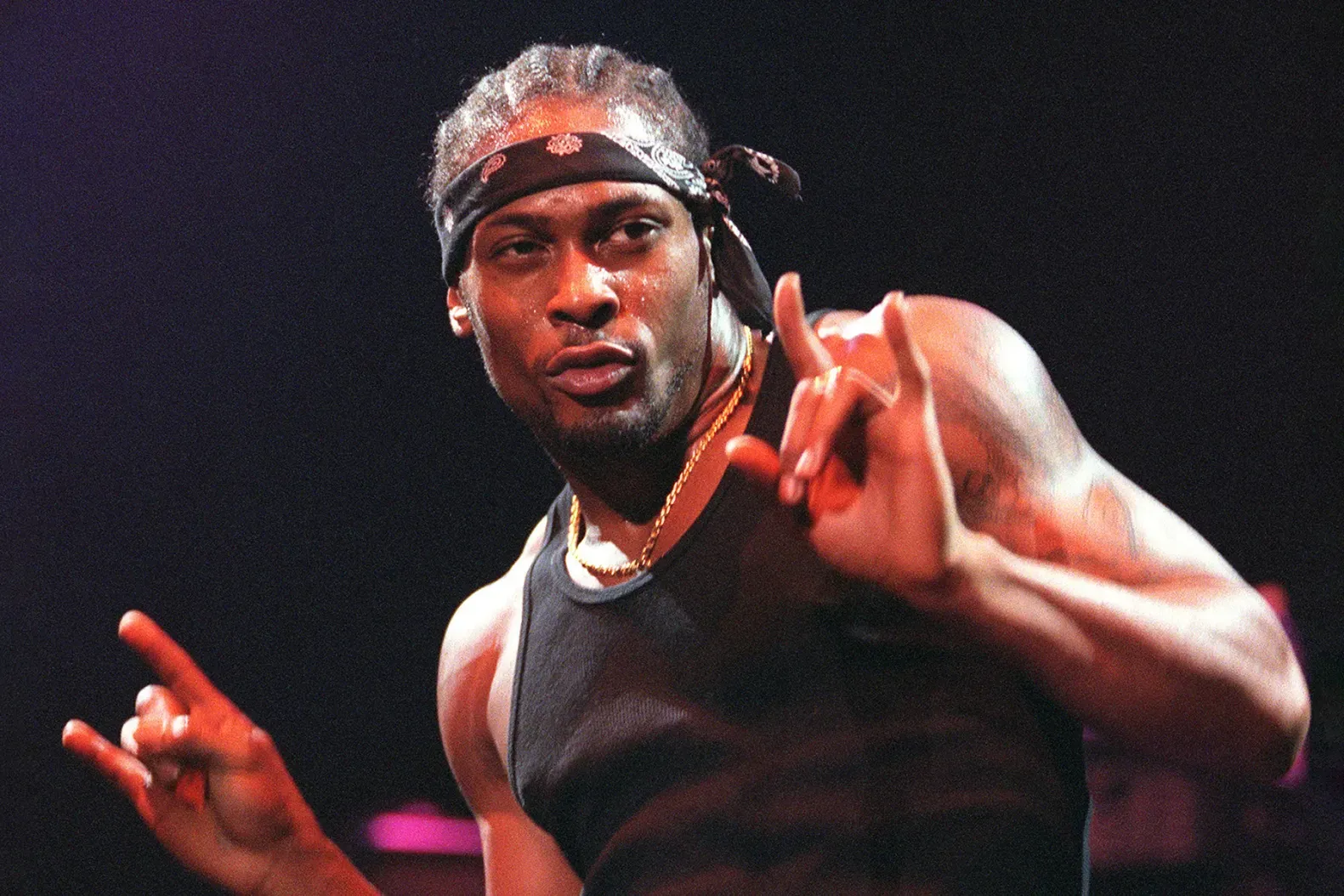 Performer in a black tank top and headband making hand gestures on a stage under red lighting