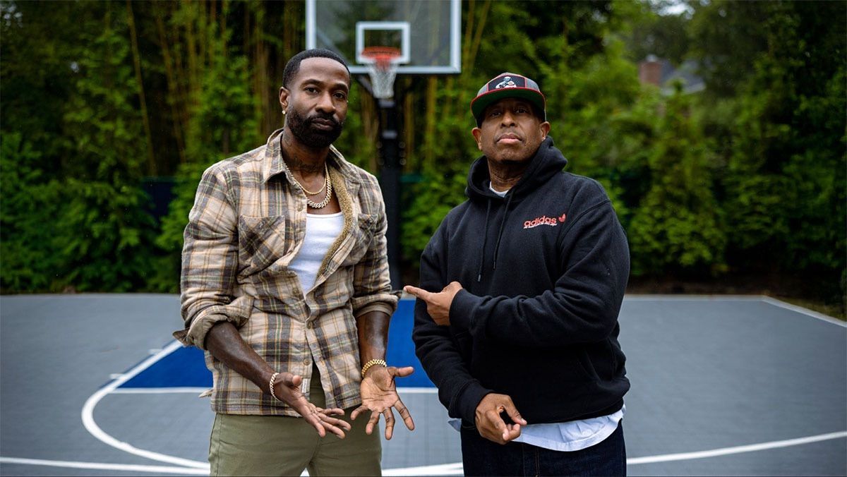 Two people standing on an outdoor basketball court, with a hoop and trees in the background.