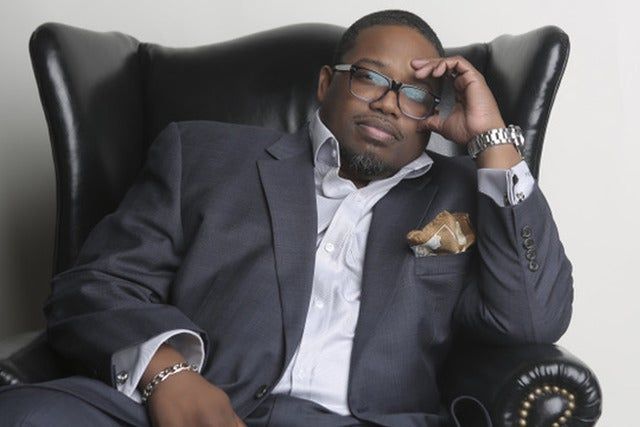 Man reclining in a black leather chair, wearing a dark suit and glasses, hand resting on his forehead.