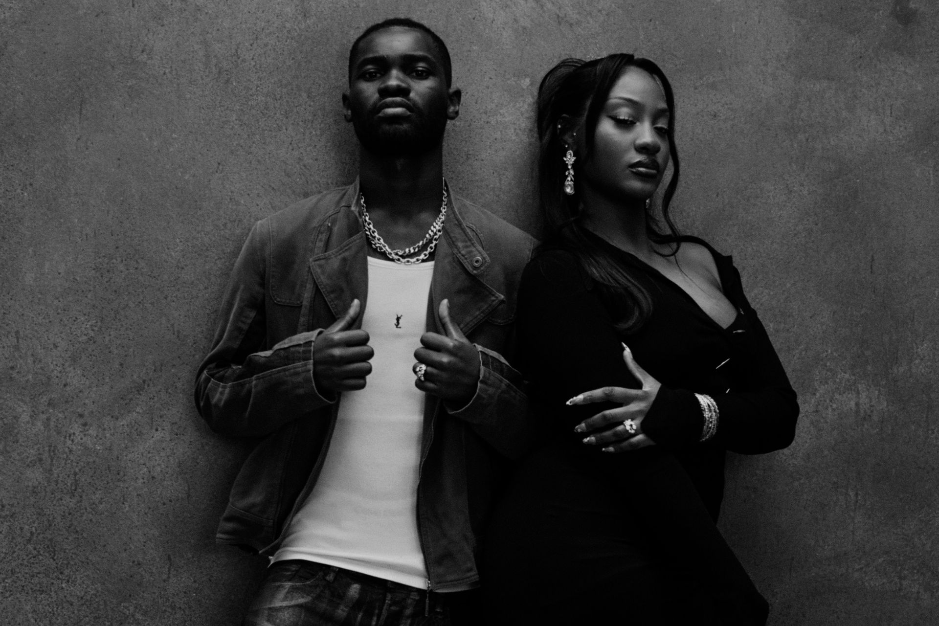 Two people posing back-to-back against a textured wall, one in a jacket and one in dark clothing.