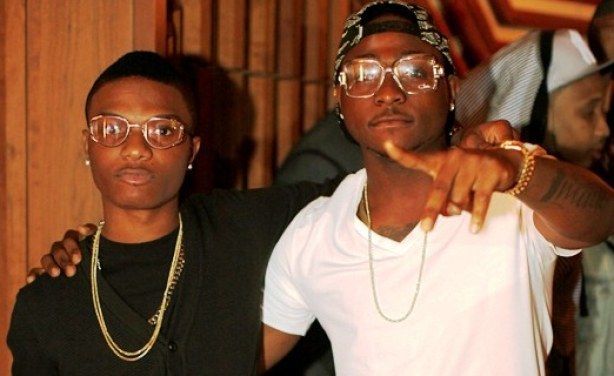 Two men posing together at an indoor event, one pointing at the camera and both wearing glasses and chains.