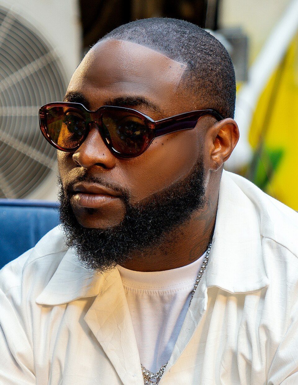 Man wearing sunglasses and a white shirt, seated indoors in a close-up portrait