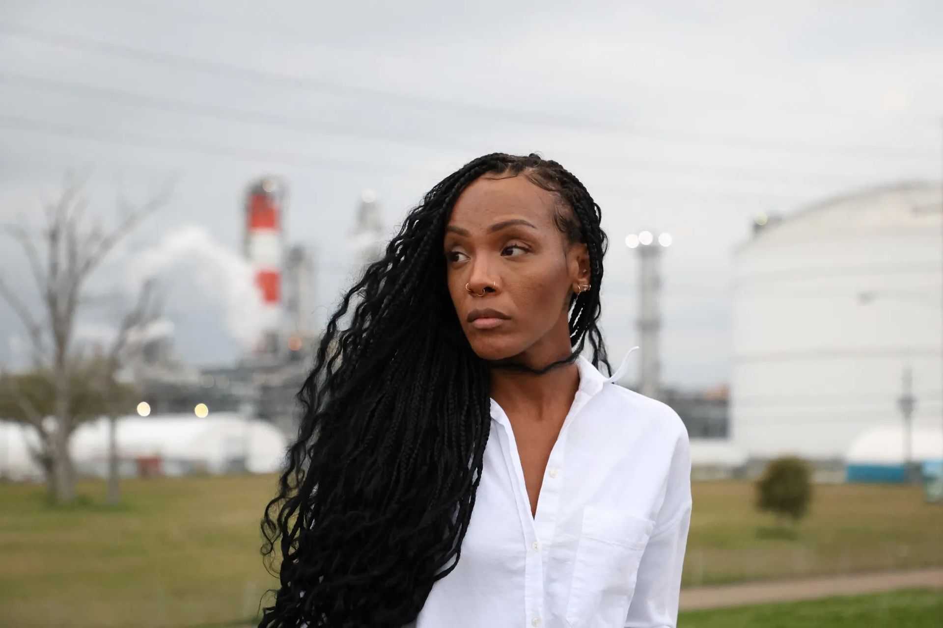 Woman with long black braids in a white shirt standing outdoors near industrial tanks under a cloudy sky