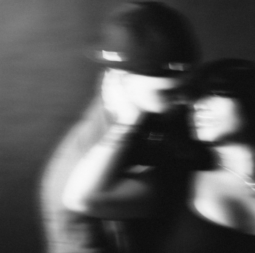 Blurry black-and-white close-up of two people embracing, one wearing a cap.