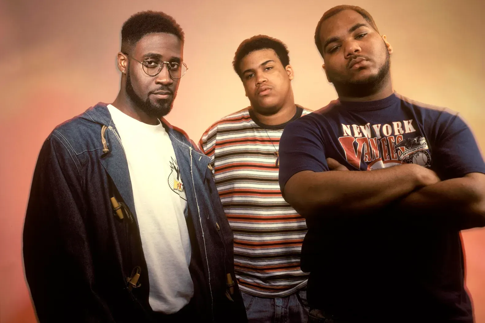 Three men posing against a warm pink backdrop, with casual clothes and serious expressions.