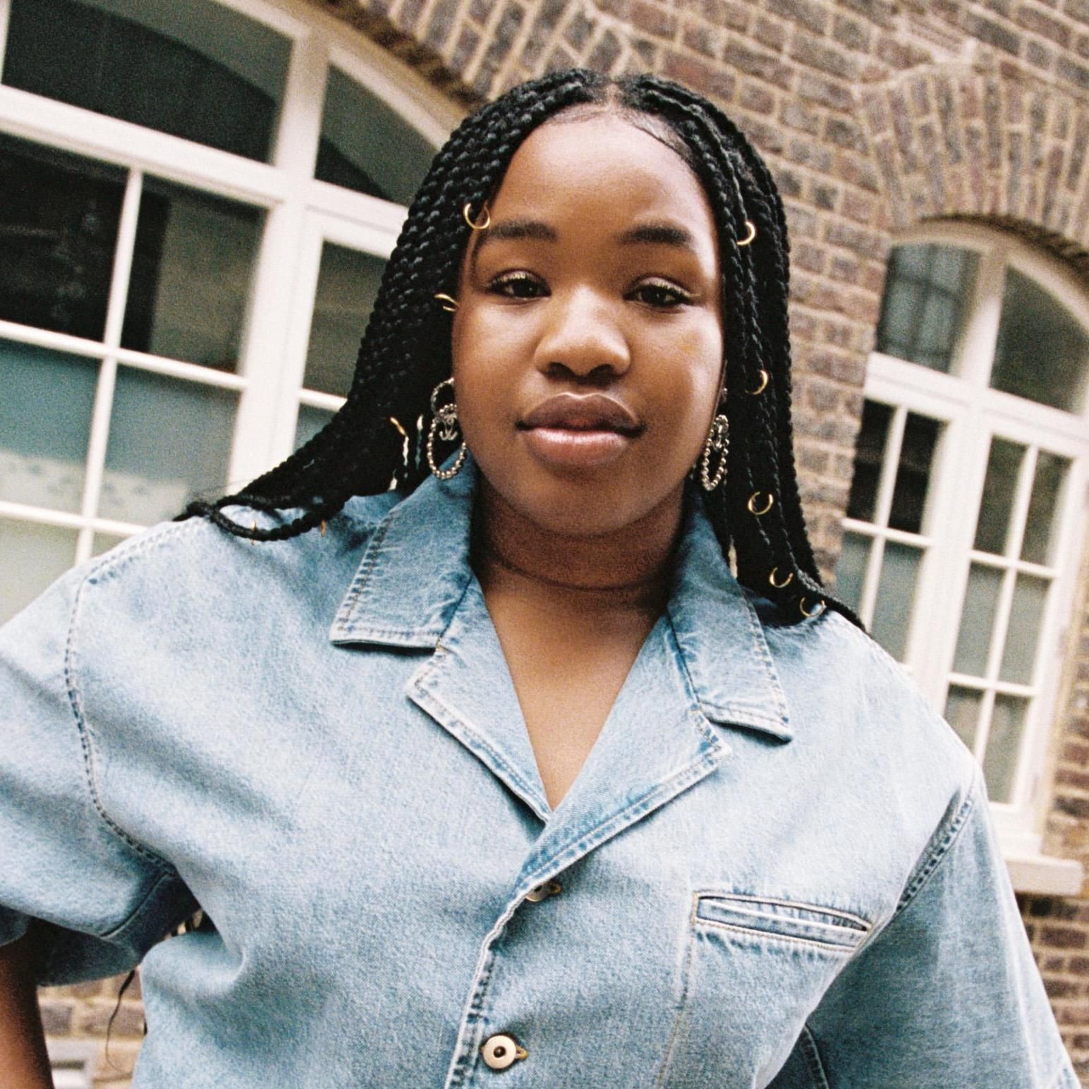 Person in a light denim jacket standing outdoors in front of large windows.
