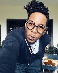 Person leaning toward the camera in glasses and a dark jacket, indoors by a window and plant.