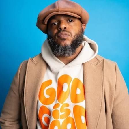Man in tan hat and coat over white hoodie with orange lettering, standing against a blue background