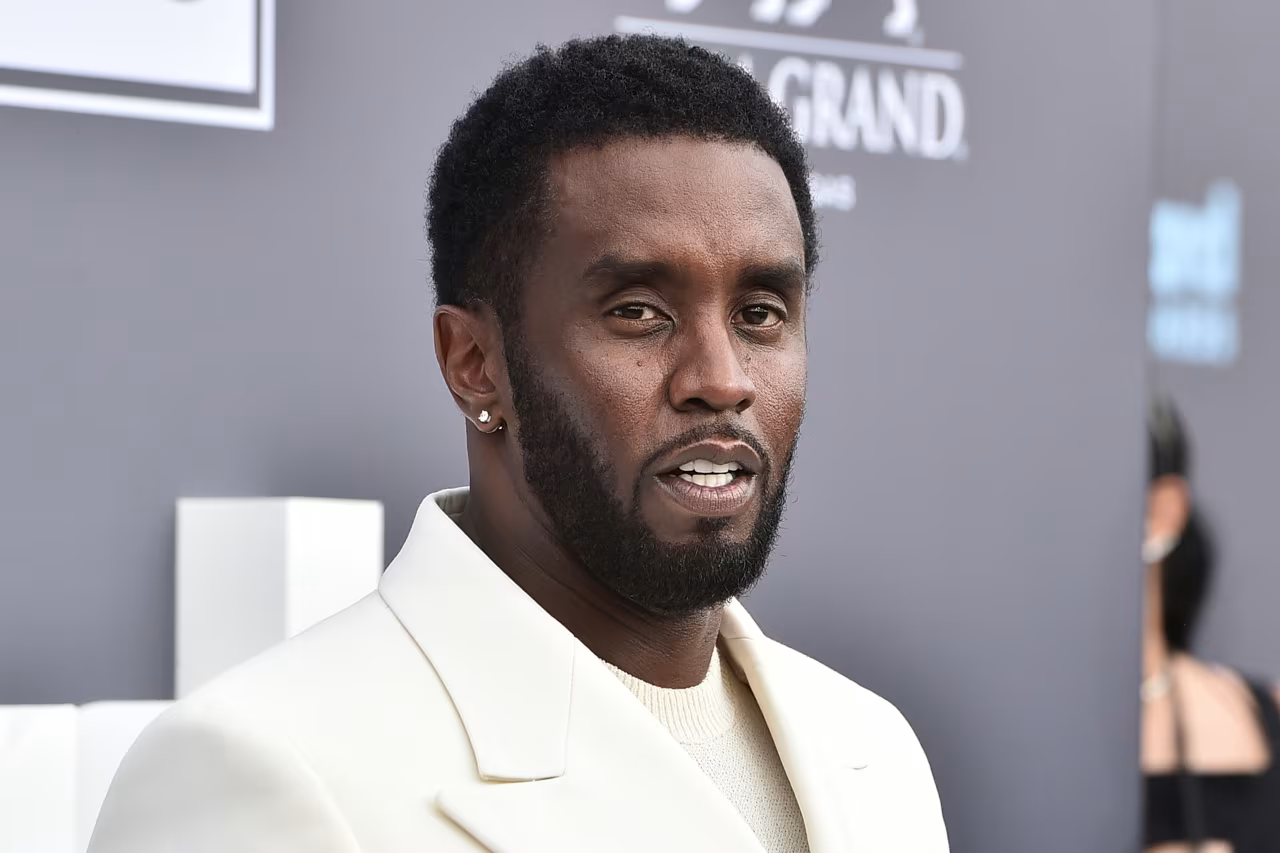 Man in a white suit posing on a red carpet with a neutral expression