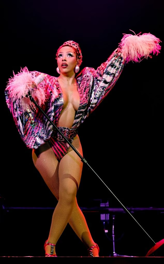 Performer in a pink, sequined stage costume with feathered sleeves, posing under spotlight on a dark stage