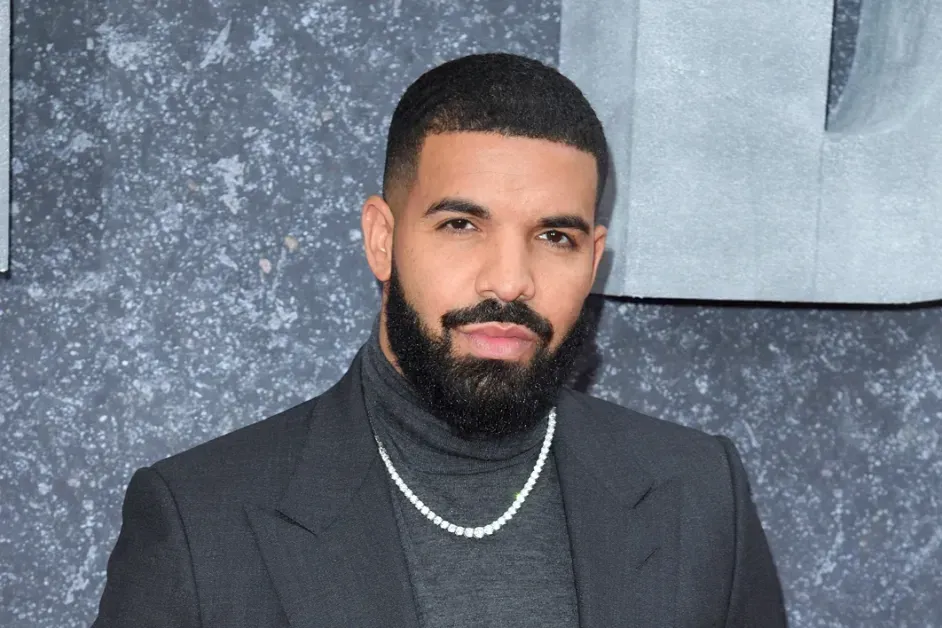 Man in a black turtleneck and blazer with a silver chain, posing against a gray backdrop.
