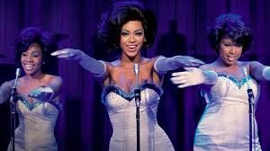 Three singers in sparkling blue stage costumes perform under purple lighting with arms outstretched.