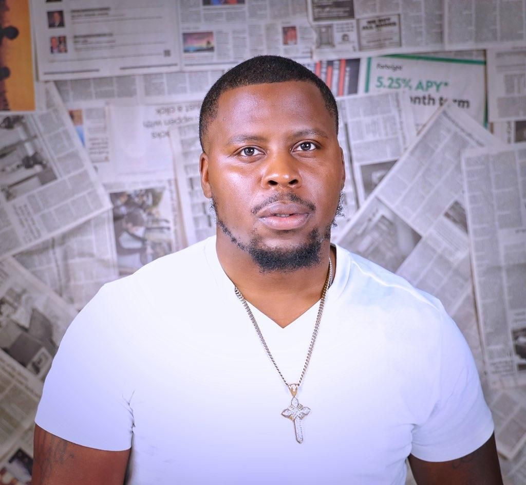 Man in a white T-shirt and cross necklace standing before a newspaper-covered wall, facing forward.