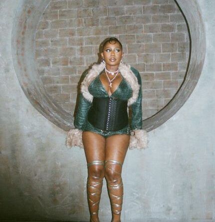 Person in a green outfit with fur trim standing against a brick wall in front of a large circular opening
