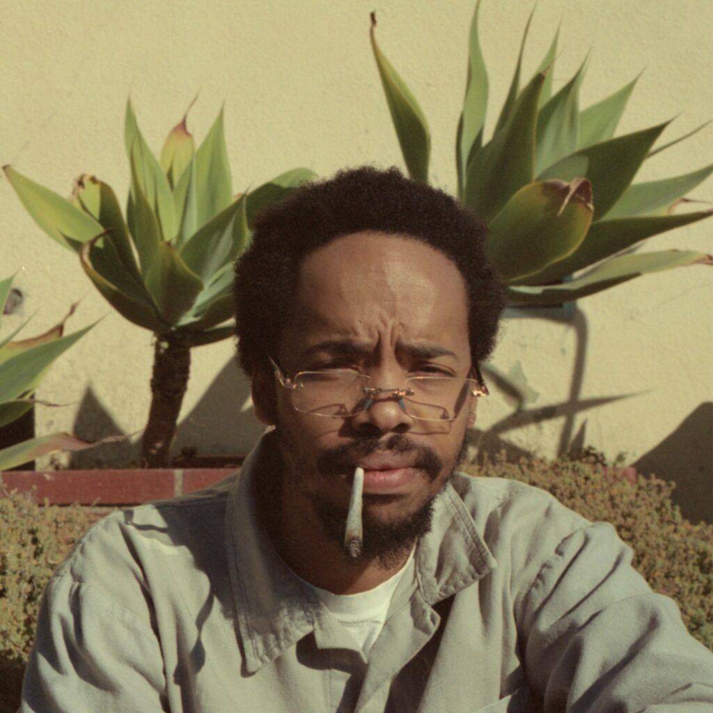 Man wearing glasses and a gray jacket, seated outdoors with large agave plants behind him.