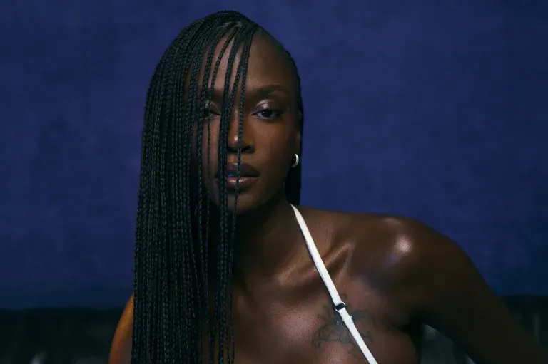 Portrait of a woman with long braids against a dark blue background, wearing a white strap top