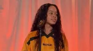 Woman in orange jersey speaking at a microphone against a red-lit backdrop