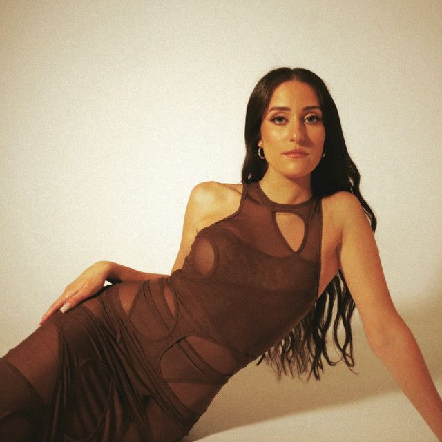 Woman reclining in a sheer brown dress against a plain beige backdrop.