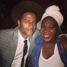 Two smiling people pose together indoors, one in a plaid jacket and hat, the other in a white top and head wrap.