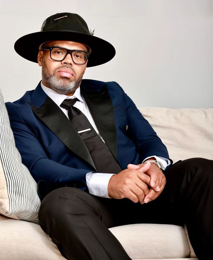 Man in a navy suit and black hat sitting on a sofa, wearing glasses and a black tie.