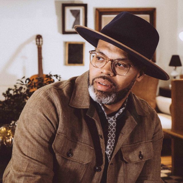 Man in a black hat and glasses sitting indoors, looking to the side in a warmly lit room.