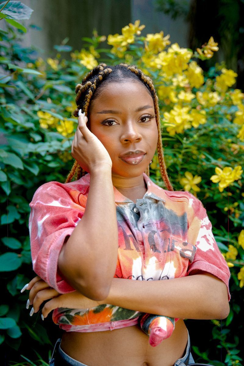 Person standing outdoors in a pink tie-dye shirt, hand to face, with yellow flowers behind them