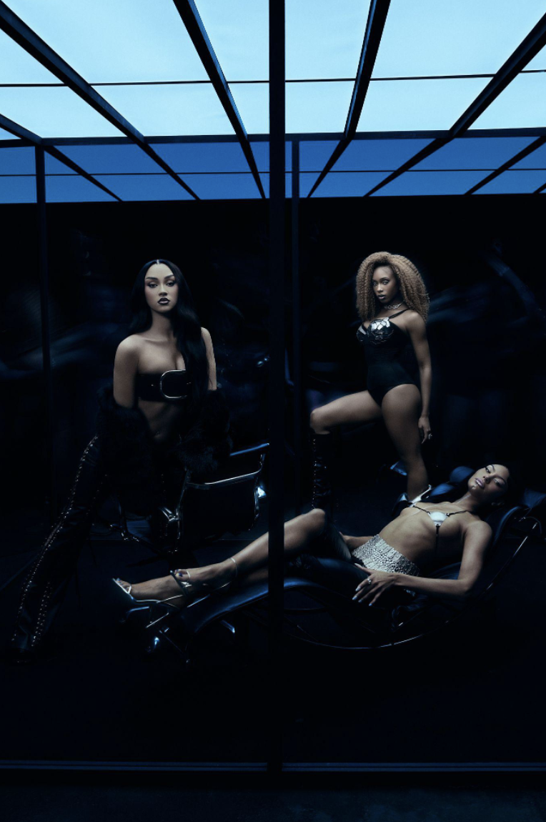 Three women in black outfits posed in a dark room with a blue skylight above.