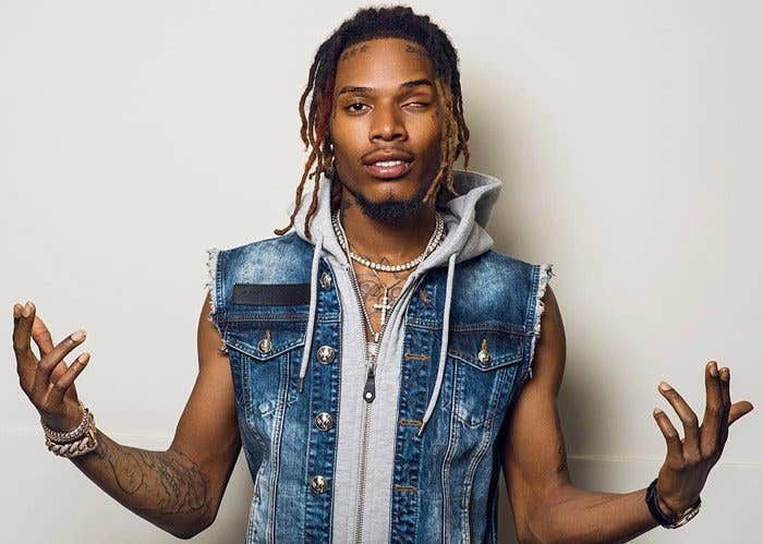 Person in a sleeveless denim vest with layered jewelry, standing against a plain background with hands raised.