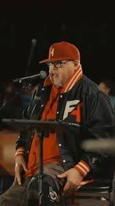 Man in red cap and black-orange jacket speaking at a microphone on a dimly lit stage