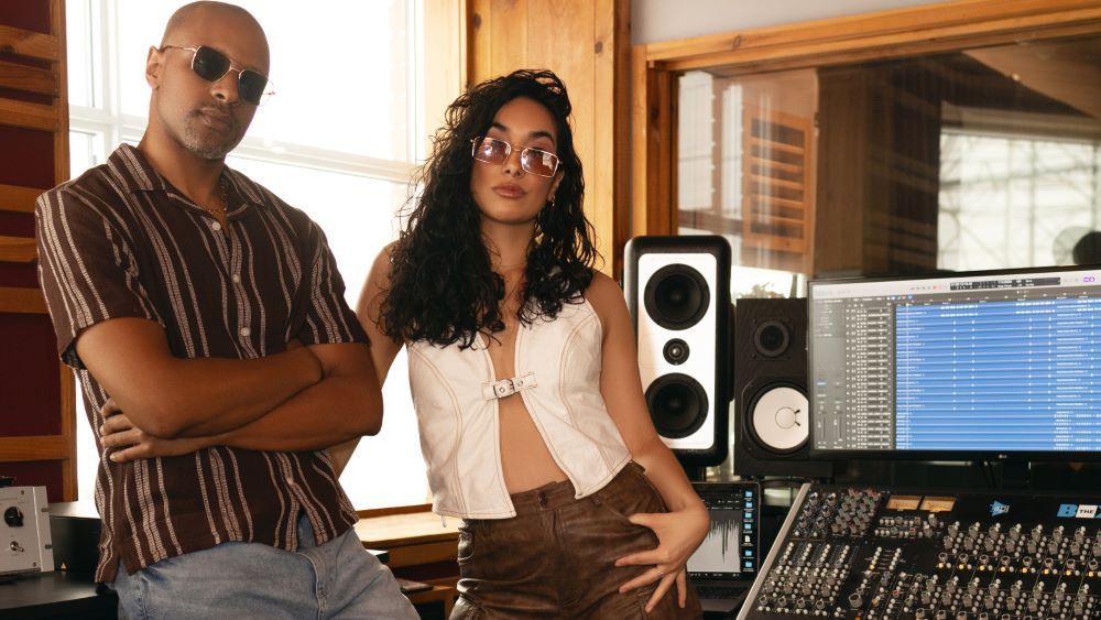Two people posing in a recording studio beside speakers and a mixing console