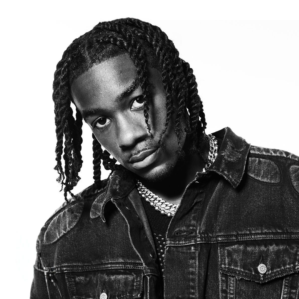 Black-and-white portrait of a person with braided hair, a denim jacket, and a chain necklace, gazing at the camera