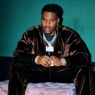 Person in a dark velvet outfit and silver chains sits on a tufted couch against a teal background.