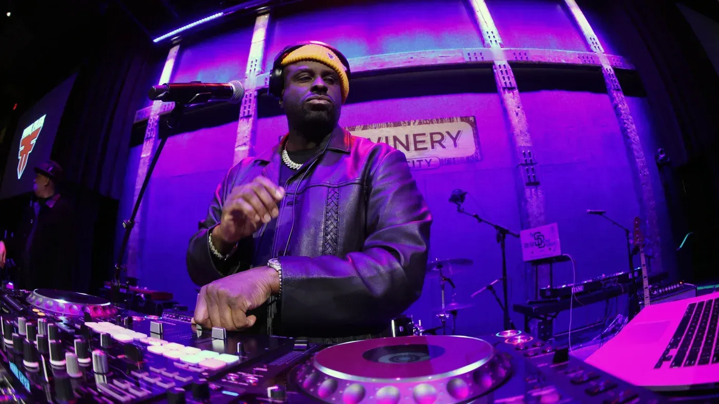 DJ performing at a purple-lit club booth, hand raised over a mixing console and laptop.