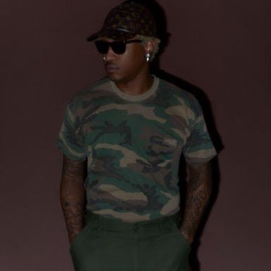 Person in sunglasses and camouflage shirt standing against a dark wall.