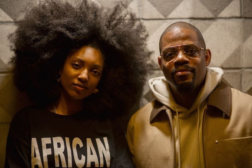Two people standing in front of a geometric wall, one in a black “AFRICAN” shirt and one in a tan jacket.