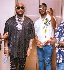 Two men posing indoors; one in black sunglasses and chains, the other in a white shirt with hand raised.