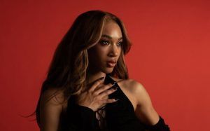 Woman with long wavy hair posing against a red background, wearing a black top and looking to the side.
