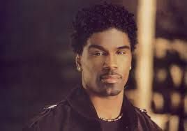 Close-up portrait of a man with curly hair and a trimmed goatee, wearing a dark jacket and chain necklace.