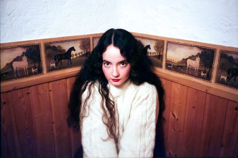 Woman with long dark hair in a white sweater, seated against a wood-paneled wall with vintage mural trim.