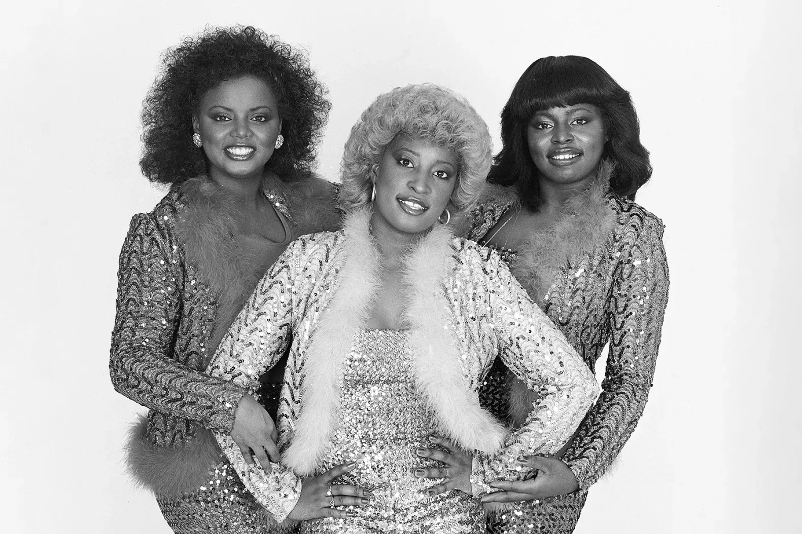 Three women posing together in sparkly outfits against a plain background