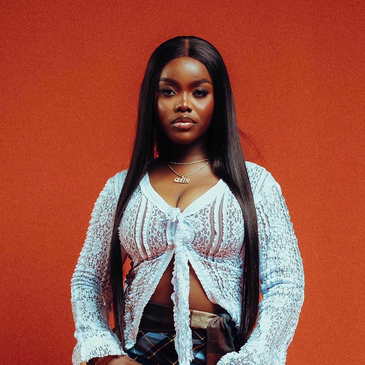 Woman standing against an orange background, wearing a white knit top and black pants.