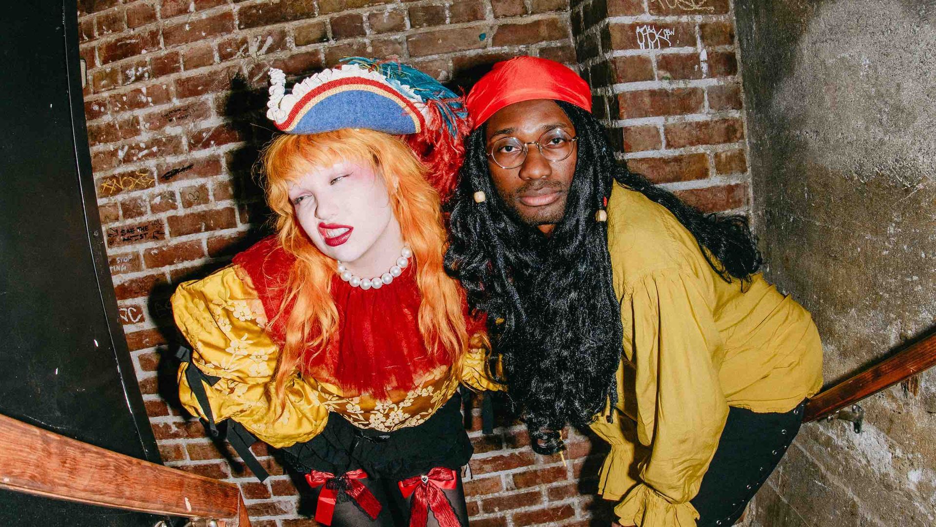 Two people in colorful costumes and helmets pose against a brick wall.