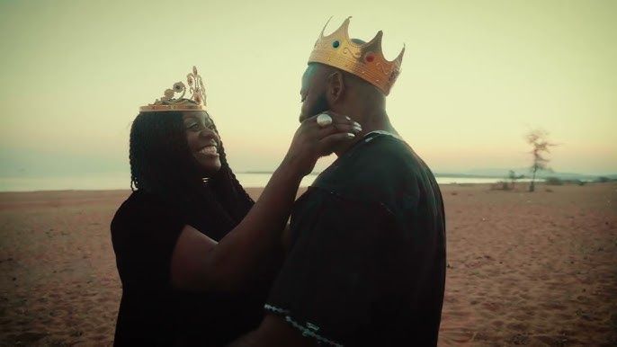 Two people in paper crowns stand on a beach at sunset, facing each other and touching faces.